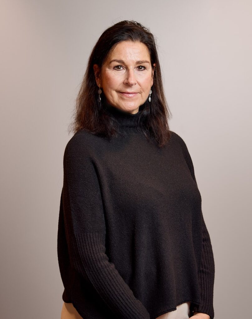 Eine Frau mit dunklem Haar trägt einen schwarzen Pullover und lächelt in die Kamera.